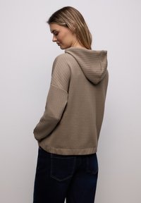 Femme aux cheveux blonds mi-longs portant un sweat à capuche beige texturé et un jean bleu foncé, regardant vers le bas avec les mains dans les poches.