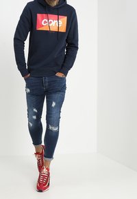 Sudadera de navy con un gráfico rojo y naranja del logo "core", combinada con jeans azules desgastados y zapatillas rojas con cordones blancos.