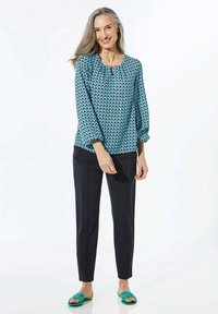 Blusa a fantasia blu con forme geometriche, abbinata a pantaloni neri e sandali verde scuro. La blusa presenta un collo a chiave e maniche morbide.