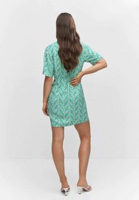 Femme aux longs cheveux bruns portant une robe courte verte et blanche à motif de feuilles et des sandales à talons blancs, se tenant avec une main sur le dos.