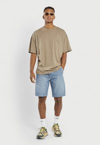 Beige oversized t-shirt with a pocket, paired with light blue denim shorts. Model wears sunglasses and beige sneakers with black accents.