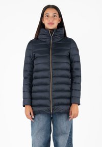 Marine puffer jacket met hoge kraag, quilted ontwerp, ritsluiting en beige accenten. Draagt over lichtblauwe denim.