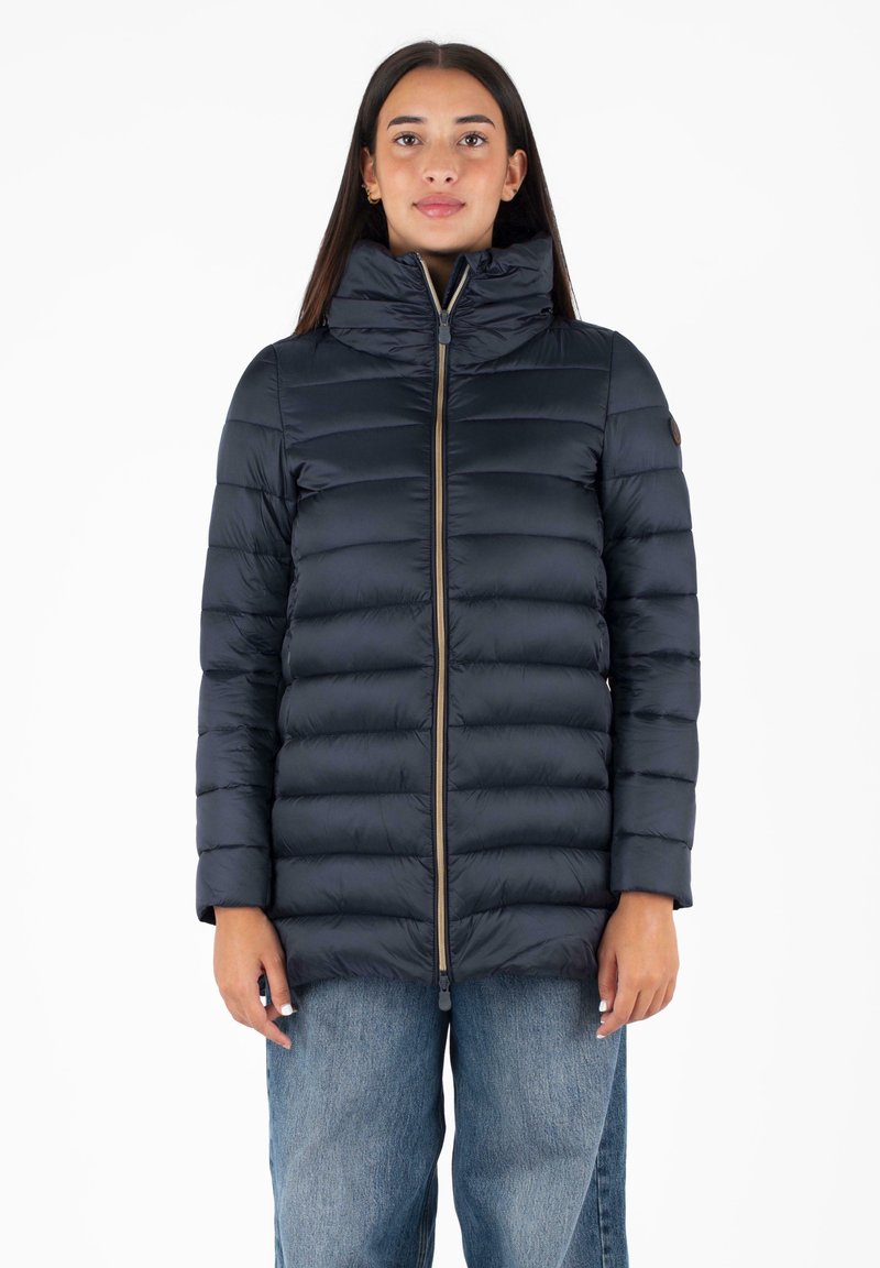 Marine puffer jacket met hoge kraag, quilted ontwerp, ritsluiting en beige accenten. Draagt over lichtblauwe denim.