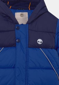 Chaqueta acolchada en tonos de azul con sección superior en azul marino, botones a presión y logo blanco; cuenta con ribetes grises y un suave forro en el cuello.
