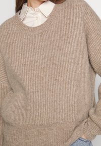 Persona che indossa un maglione a coste beige sopra una camicia bianca con il colletto, con una mano infilata nella tasca dei jeans blu.