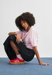 Rosa kort T-skjorte, svarte vide bukser og røde joggesko med hvite striper. Modell sittende på et blått teppe, hår i naturlige krøller.