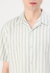 Gaišzaļš un balts īsajām piedurknēm krekls ar apkakli, ar vertikālām svītrām. Izgatavots no teksturēta auduma, ar pieciem pogām priekšpusē.