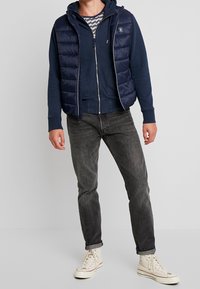 Chaleco acolchado azul marino sobre una sudadera azul marino con una camiseta a rayas, combinado con jeans gris oscuro y zapatillas altas blancas.