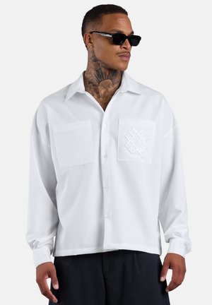 Mann mit Hals-Tattoos, der schwarze Sonnenbrille und ein weißes Hemd mit zwei Brusttaschen trägt, stehend vor einem einfarbigen Hintergrund.