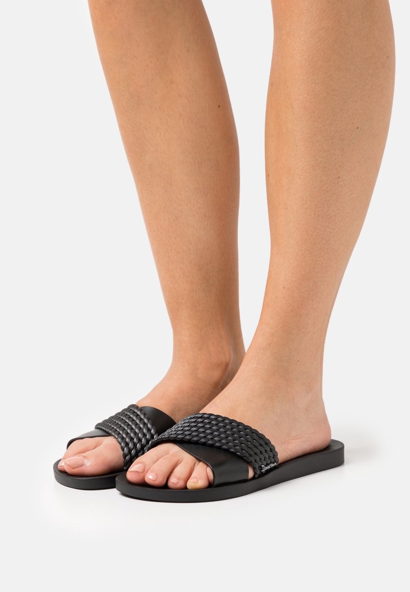 Pieds portant des sandales à lanière tissée noire avec semelles plates sur un fond neutre.