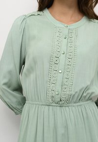 Hellgrünes langärmliges Kleid mit Spitzenborte entlang der Knopfleiste, runden Knöpfen, geraffter Taille und filigranen Spitzelementen an den Schultern.