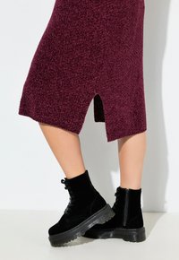 Stivaletti in suede neri con suola trasparente spessa, lacci sulla parte anteriore e zip laterale. L'outfit è completato da un vestito a maglia bordeaux con uno spacco.
