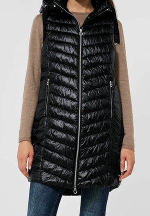 Gilet rembourré noir avec fermeture éclair sur le devant, design matelassé en chevrons, capuche et deux poches latérales, porté par-dessus un pull beige et un jean bleu.