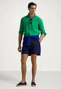 Zielona lniana koszula button-up, granatowe szorty, niebieski sweter przewiązany w pasie, okulary przeciwsłoneczne i niebieskie espadryle. Mocny, swobodny strój na lato.