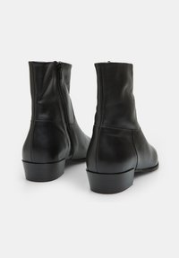 Botas de tobillo de cuero negro con una textura suave, puntera redondeada y cremallera lateral. Presentan un tacón bajo apilado. Diseño limpio con acentos mínimos.