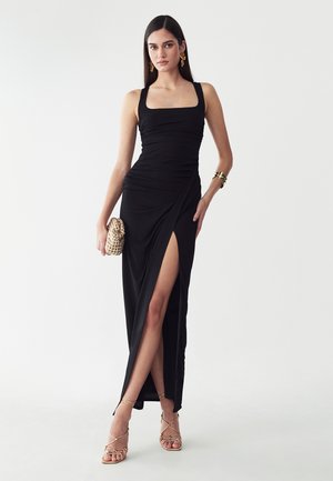 Donna che indossa un vestito nero senza maniche con uno spacco alto, sandali dorati, orecchini dorati, braccialetto e tiene una piccola pochette dorata.