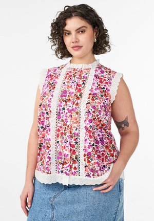 Kvinde med krøllet hår iført en ærmeløs blomstret bluse med blonder og en blå denim nederdel, som viser en katteansigt-tatovering på sin arm.