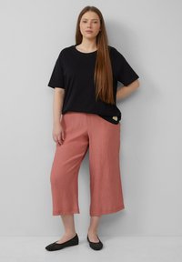 T-shirt noir en coton à manches courtes, associé à un pantalon large plissé rose clair. Chaussures plates noires avec une surface texturée.