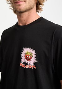 Camiseta negra de algodón con un diseño gráfico colorido que presenta una cara de estilo caricaturesco y el texto "VOLCOM" en la parte inferior. Textura suave, cuello redondo.