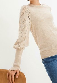 Beige stickad tröja med texturerat öppet arbete och upphöjda prickmönster på puffiga ärmar, kombinerad med blå jeans, hand vilande på baksidan av en trästol.