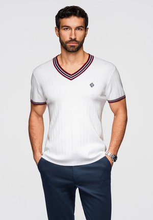 Man met kort donker haar en baard, gekleed in een witte V-hals trui met korte mouwen met gestreepte bies en een donkerblauwe broek, handen in de zakken.