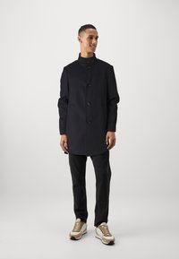 Marineblauwe jas met een hoge kraag en vijf knopen, gecombineerd met zwarte jeans en beige sportschoenen met witte en zwarte accenten.