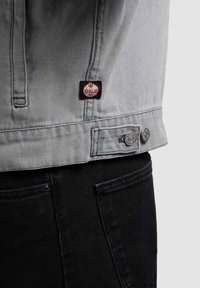 Ourlet de veste en jean gris avec un patch logo Khmer noir et rouge sur un pantalon en jean noir, montrant la poche arrière et les détails des coutures.