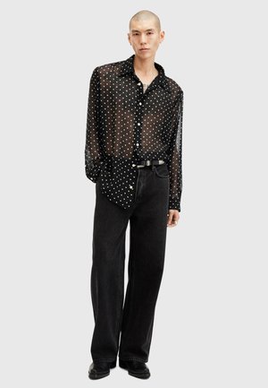 Camicia nera trasparente con bottoni e pois bianchi, abbinata a jeans neri a vita alta. Tessuto morbido, vestibilità rilassata e maniche lunghe.