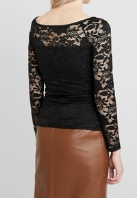 Haut en dentelle noire à manches longues, présentant des motifs floraux et un décolleté en forme de U. Le matériau est délicat et texturé, associé à une jupe en cuir marron.
