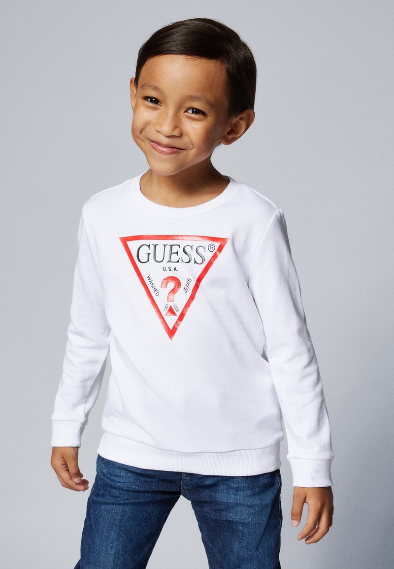 Felpa bianca a maniche lunghe con un logo triangolare rosso raffigurante "GUESS U.S.A." e un motivo grafico, abbinata a jeans in denim blu.