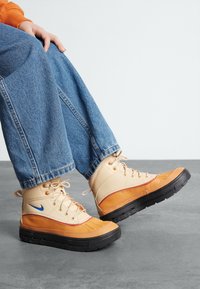 High-top sneakers featuring a tan leather upper with orange accents, a blue logo, and a thick black rubber sole. Denim jeans partially visible.