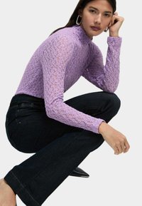 Maglia lunga a maniche con tessuto di pizzo testurizzato di colore viola chiaro, abbinata a jeans a campana in denim scuro, che evidenziano una silhouette aderente.