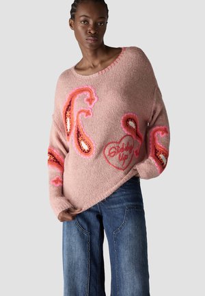 Woman with braided hair wearing a pink sweater with orange paisley designs and the words "Baby Up" paired with blue jeans against a plain background.