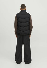 Schwarze Steppweste mit hohem Kragen, ausgestattet mit einem Chevron-Steppmuster und leichtem Material, getragen über einem dunkelbraunen Langarmshirt.