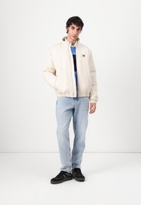 Beige bomberjakke med høj krave, elastiske manchetter og kant, kombineret med lyseblå jeans og sorte sneakers. Lille logo detalje på brystet.
