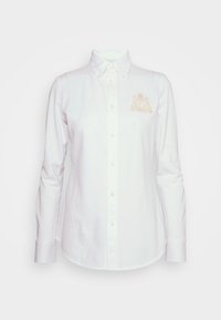 Vit skjorta med knappar gjord av bomull, med långärmar, en spetsig krage och en broderad guld-logotyp på vänster bröst.