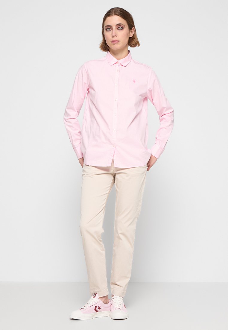 Chemise boutonnée rose clair à manches longues, associée à un pantalon beige et des baskets roses, avec un petit logo sur la chemise.