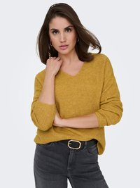 Senfgelber, gerippter Pullover mit V-Ausschnitt, lockerer Passform und hochgekrempelten Ärmeln. Getragen mit dunkelgrauen Jeans und einem schwarzen Gürtel mit goldener Schnalle.