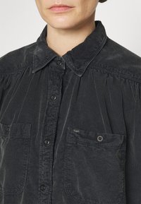 Primo piano di una persona che indossa una camicia nera in corduroy con bottoni, dotata di due tasche sul petto, una delle quali etichettata "Lee".