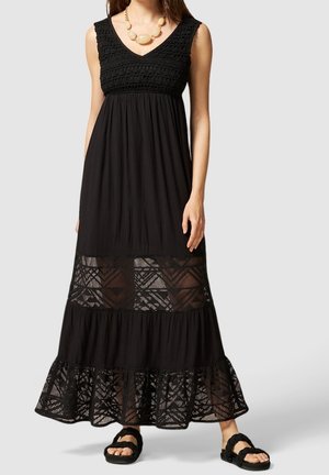 Robe maxi noire avec un corsage texturé, des détails en dentelle en couches à l'ourlet, et une coupe ample. Associée à des sandales tressées noires pour le contraste.
