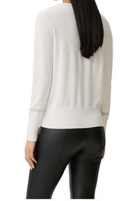 Biały sweter na drutach z długimi rękawami i okrągłym dekoltem, z subtelnymi zdobieniami na rękawach, zestawiony z czarnymi legginsami.