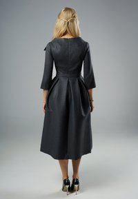 Kobieta z długimi blond włosami, ubrana w teksturowaną, ciemnoszara sukienkę midi i czarne wysokie obcasy, stojąca tyłem na jednolitym tle.