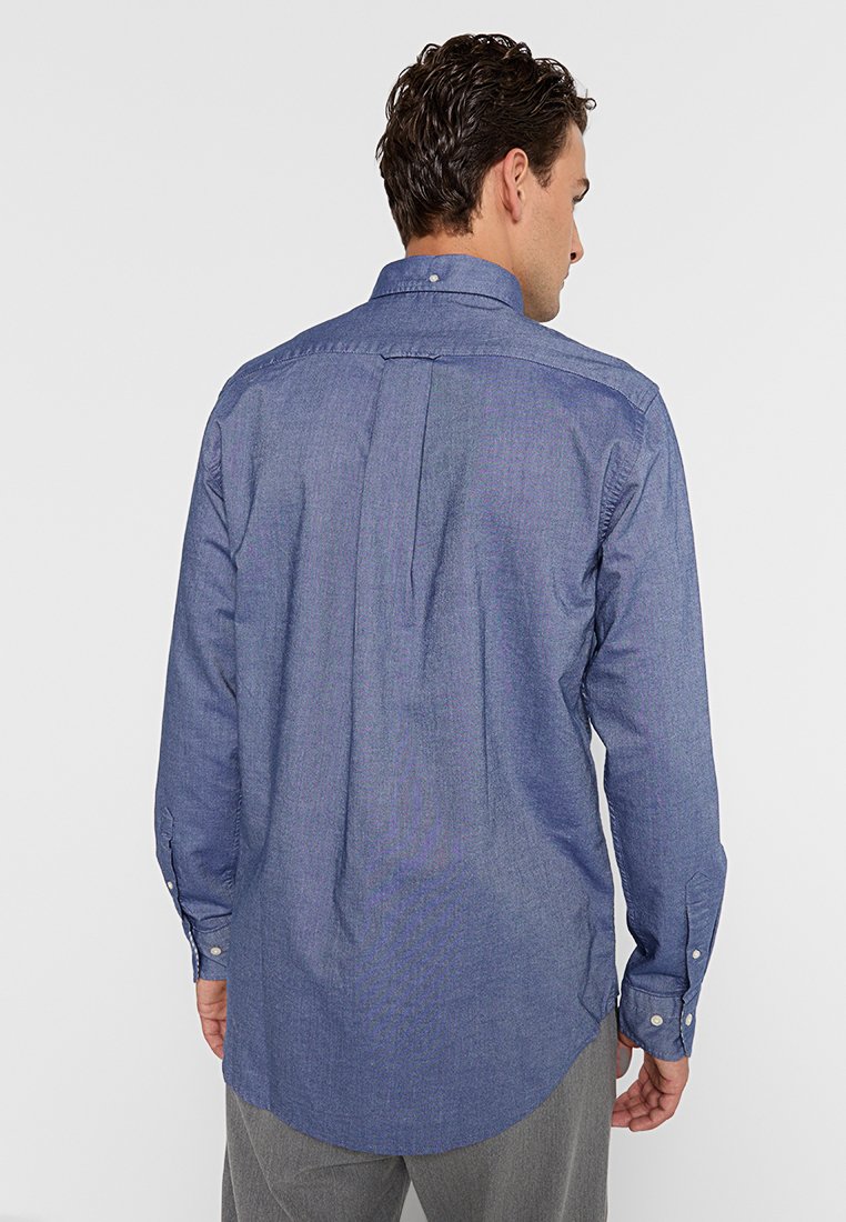 Camisa de hombre de manga larga en chambray azul oscuro. Presenta un collar abotonado, mangas con puños y un pliegue en la espalda para mayor movimiento.