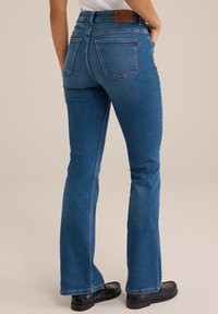 Siniset denim-mittarit, joissa on keskikorkea vyötärö, takataskut ja ruskea nahkainen brändilogo. Mato sileä rakenne ja klassinen sininen väri.