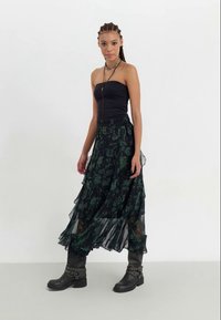 Top preto sem alças com uma saia maxi fluida e em camadas, apresentando um padrão floral verde escuro. Combinado com botas pretas robustas até o tornozelo.