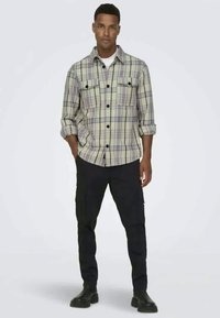 Koszula w kratę w jasnym zielonym, fioletowym i białym kolorze, z zapiętymi kieszeniami na piersi i podwiniętymi rękawami, w połączeniu z czarnymi spodniami cargo i czarnymi butami.
