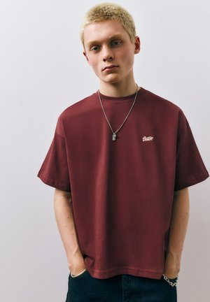 Mladá osoba s krátkymi blond vlasmi má na sebe voľné bordové tričko, strieborný náhrdelník a náramok, stojí pred jednoduchým svetlým pozadím.