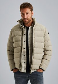 Doudoune beige avec un col montant, fermeture éclair et détails de couture, superposée sur une chemise à carreaux, portée avec un jean bleu.