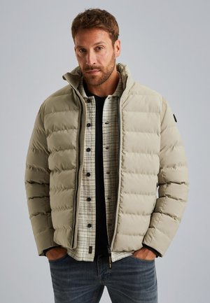 Doudoune beige avec un col montant, fermeture éclair et détails de couture, superposée sur une chemise à carreaux, portée avec un jean bleu.