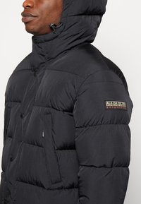 Giacca a vento nera con finitura strutturata, cappuccio rimovibile, tasche frontali e un'etichetta con logo sulla manica. Design trapuntato distintivo.